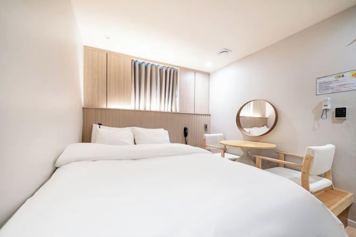 Habitación de hotel con una cama grande, una mesita pequeña, una silla, un espejo redondo y un panel de pared de madera.