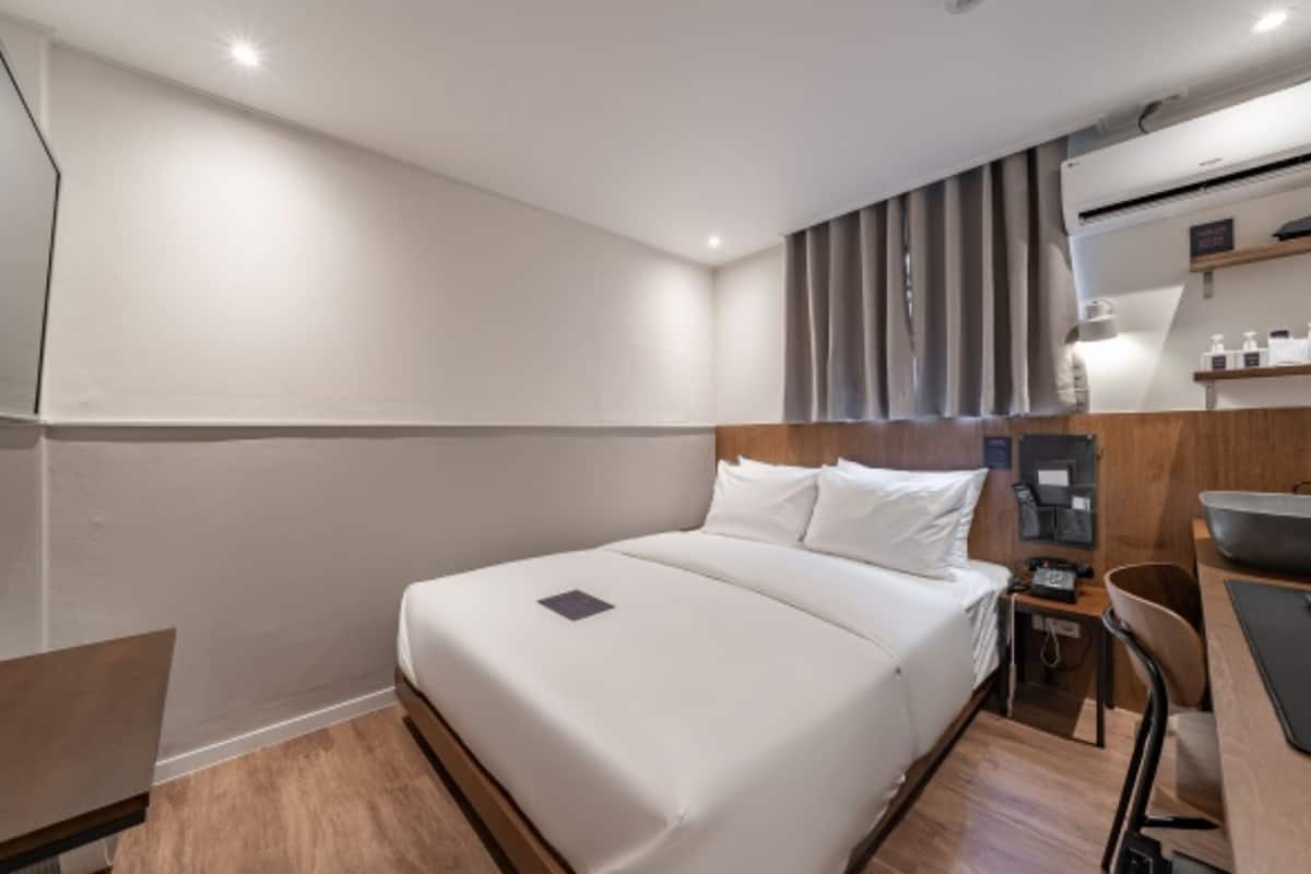 Une chambre d’hôtel moderne équipée d’un grand lit, d’un bureau, d’une chaise, d’une petite table, d’une table de chevet et d’un climatiseur fixé au mur.