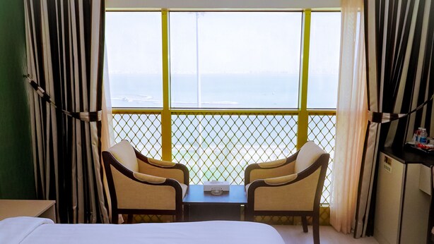 A hotel room with a bed, two chairs, a small table, and a view of the sea through a window with a yellow railing. - Capital O 126 Manama Tower Hotel (Manama)