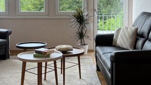 A living room with a black leather sofa, two round tables, a potted plant, and large windows offering a view of greenery.