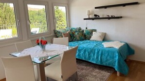 A bedroom with a bed, a table with chairs, and a window with a view of greenery.