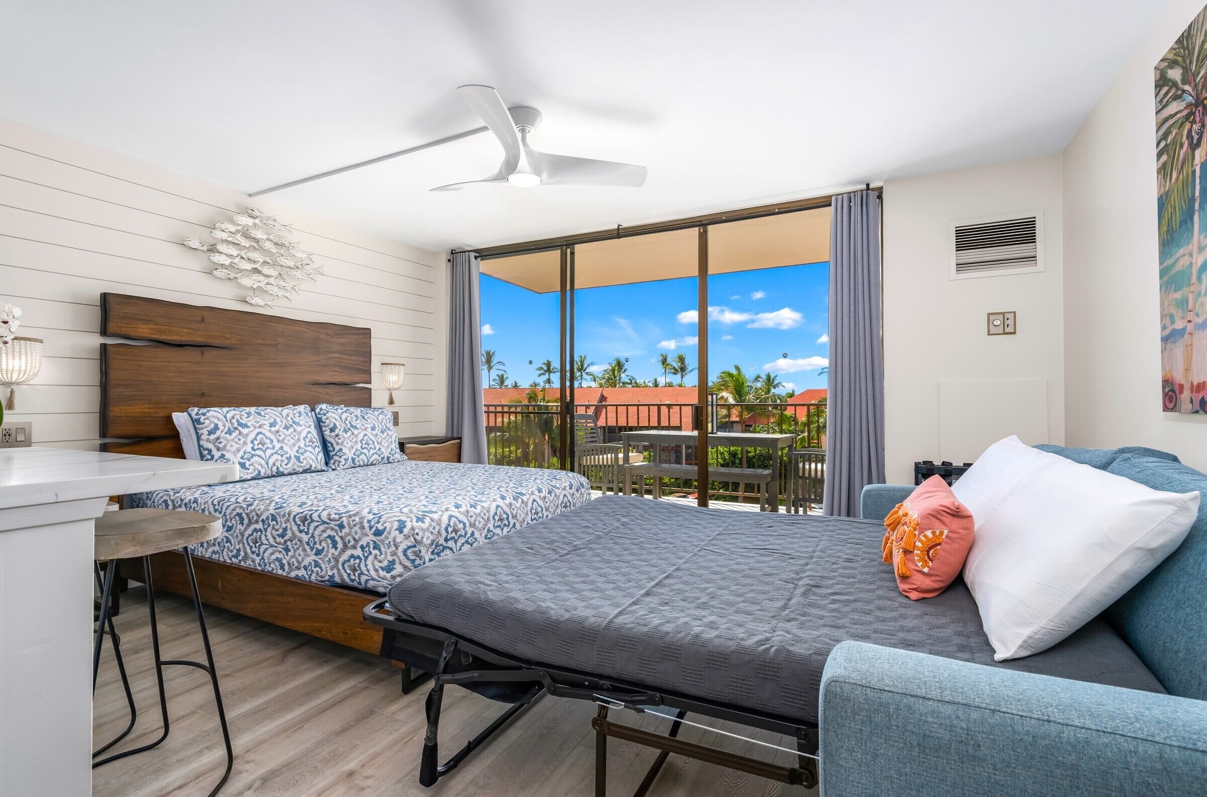Un dormitorio con dos camas, un escritorio, una silla, un ventilador de techo y un balcón con vistas a palmeras y un edificio rojo.