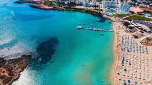 Eine Luftaufnahme einer Küstenregion mit einem Yachthafen, einem Sandstrand mit Sonnenschirmen und einem Wohngebiet mit Gebäuden.