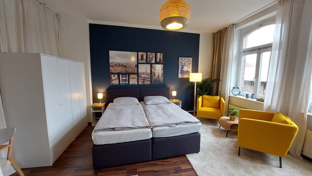 Une chambre moderne avec un grand lit, des tables de chevet, un fauteuil jaune et une fenêtre avec des rideaux.