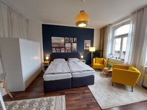 Een moderne slaapkamer met een groot bed, nachtkastjes, een gele fauteuil en een raam met gordijnen.