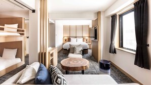 Een hotelkamer met een stapelbed, een slaapbank, een ronde houten tafel, een bed met hoofdeinde, een televisie en een raam met gordijnen.