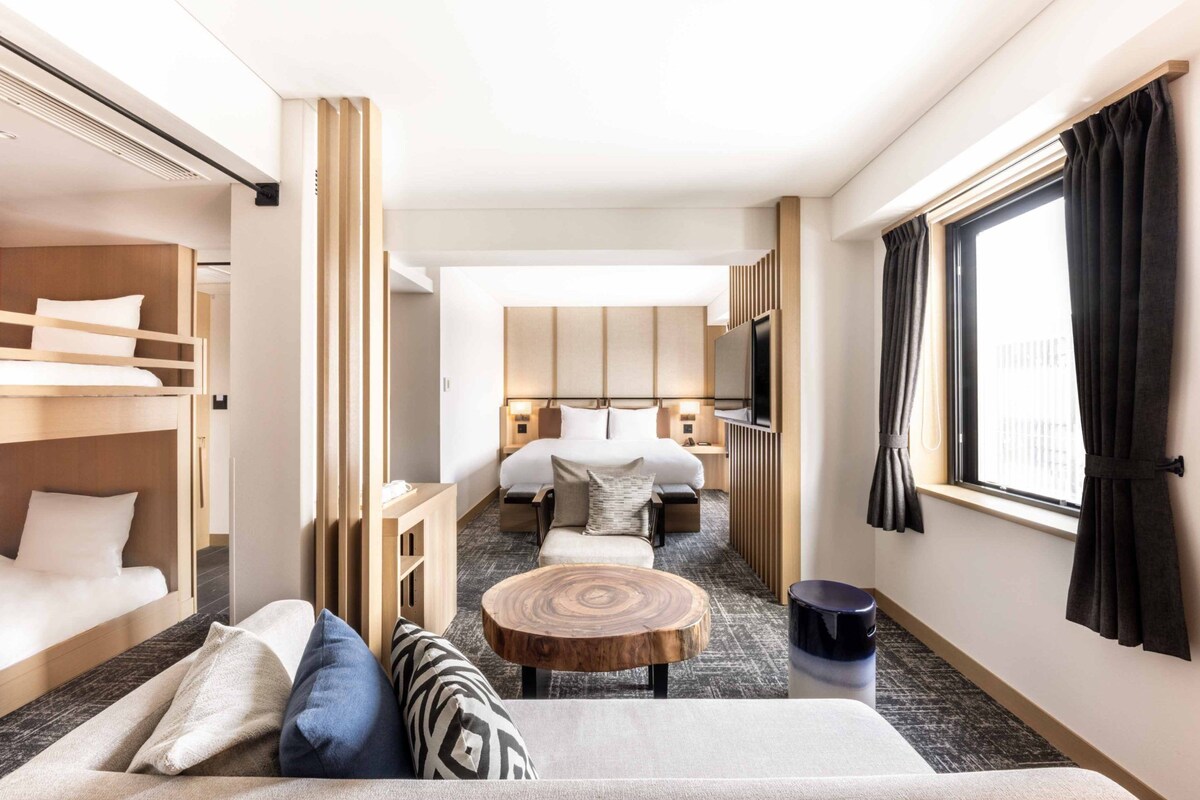 Une chambre d’hôtel avec un lit superposé, un canapé, une table ronde en bois, un lit avec une tête de lit, une télévision et une fenêtre avec des rideaux.
