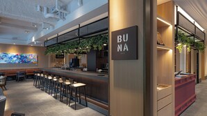 Een moderne bar met een toonbank, barstoelen en een bord waarop 'BUNA BAR' staat.