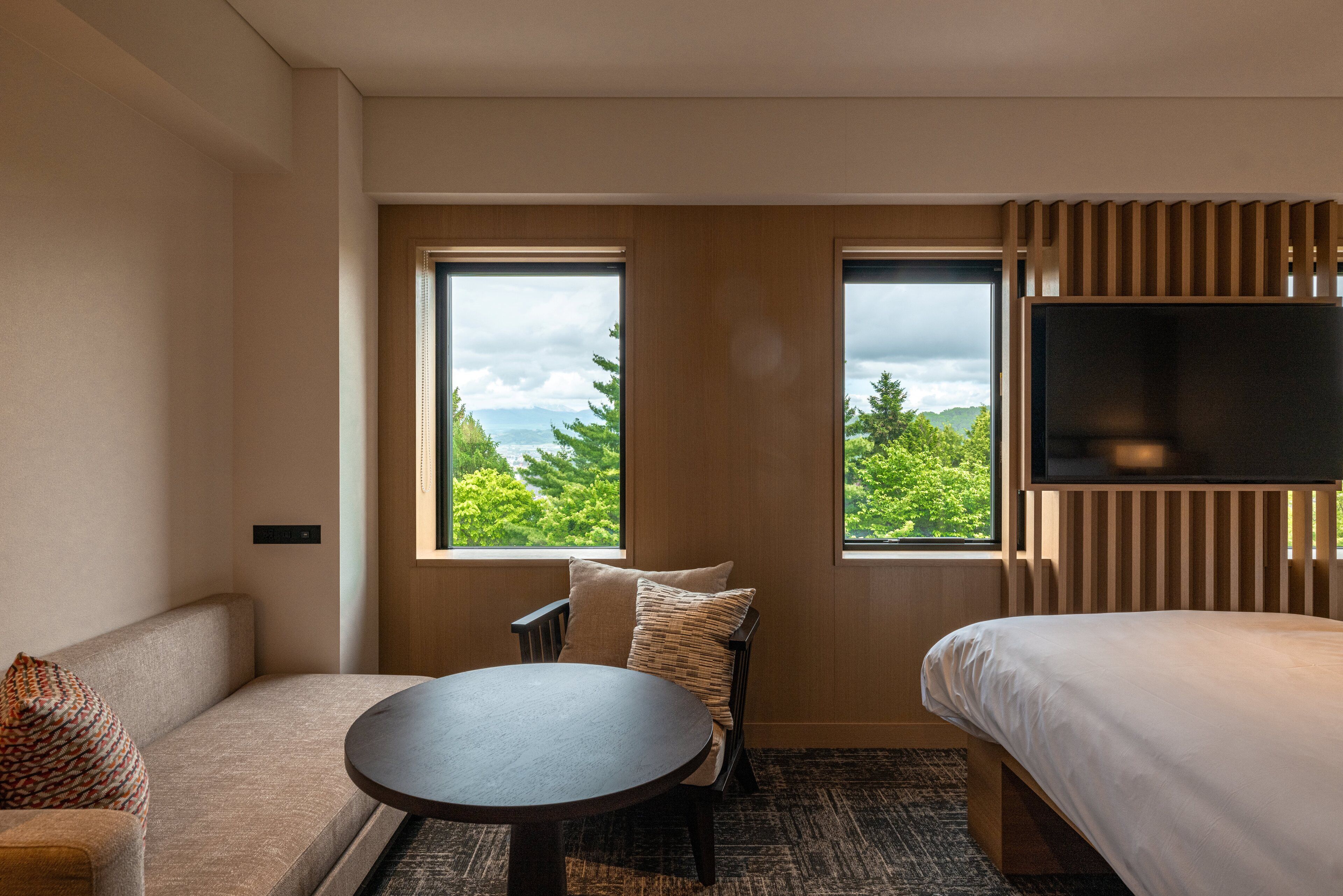 Habitación de hotel con sofá, una mesa redonda, cama y televisor. Dos ventanas ofrecen vistas a la vegetación y montañas.