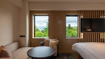 Habitación de hotel con sofá, una mesa redonda, cama y televisor. Dos ventanas ofrecen vistas a la vegetación y montañas.