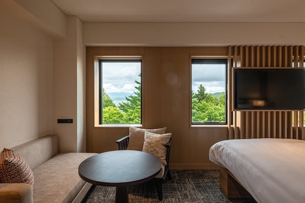 A hotel room with a sofa, a round table, a bed, and a TV. There are two windows offering a view of greenery and mountains. - Nozo Hotel (Furano)