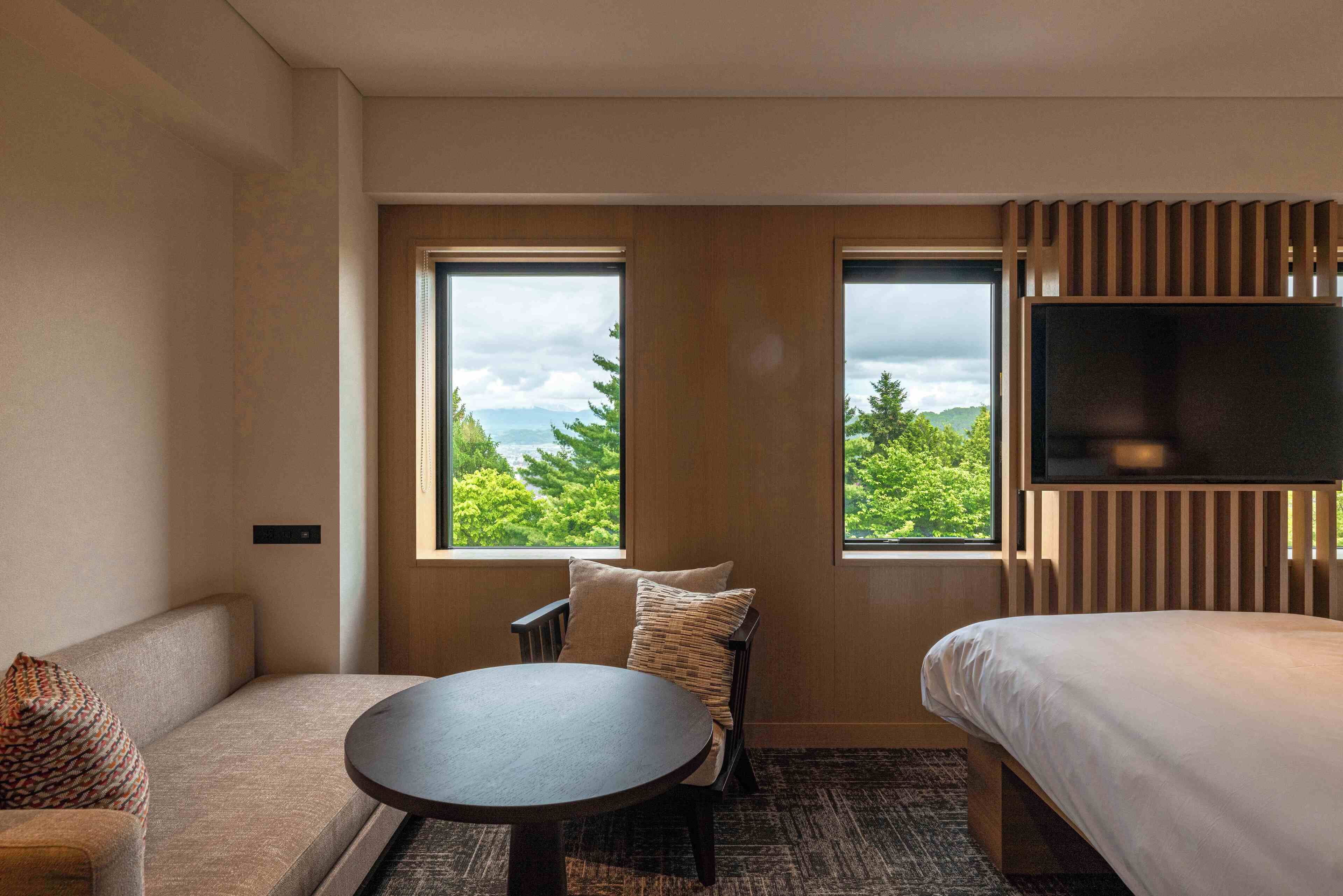 Habitación de hotel con sofá, una mesa redonda, cama y dos ventanas con vistas a zonas verdes y montañas.