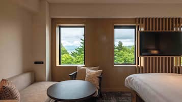 Habitación de hotel con sofá, una mesa redonda, cama y dos ventanas con vistas a zonas verdes y montañas.