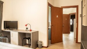 A hotel room with a bed, a desk with a television and lamp, a chair, and a trash bin. - Appartamento - Edolo (Edolo)
