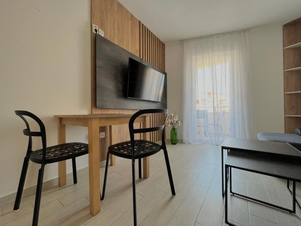 A modern living room with a flat-screen TV mounted on a wooden panel wall, two black chairs, a wooden dining table, and a large window with sheer curtains.