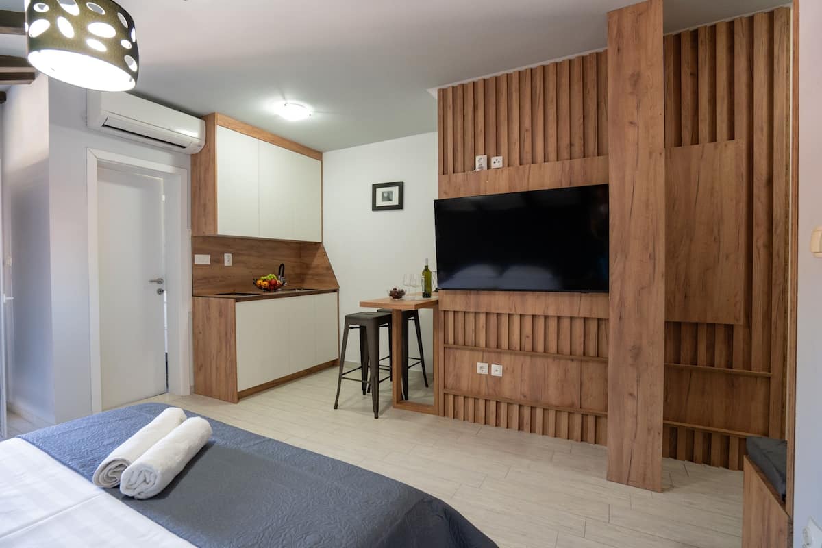 A compact hotel room with a bed, a kitchenette, a TV, and a wooden accent wall.
