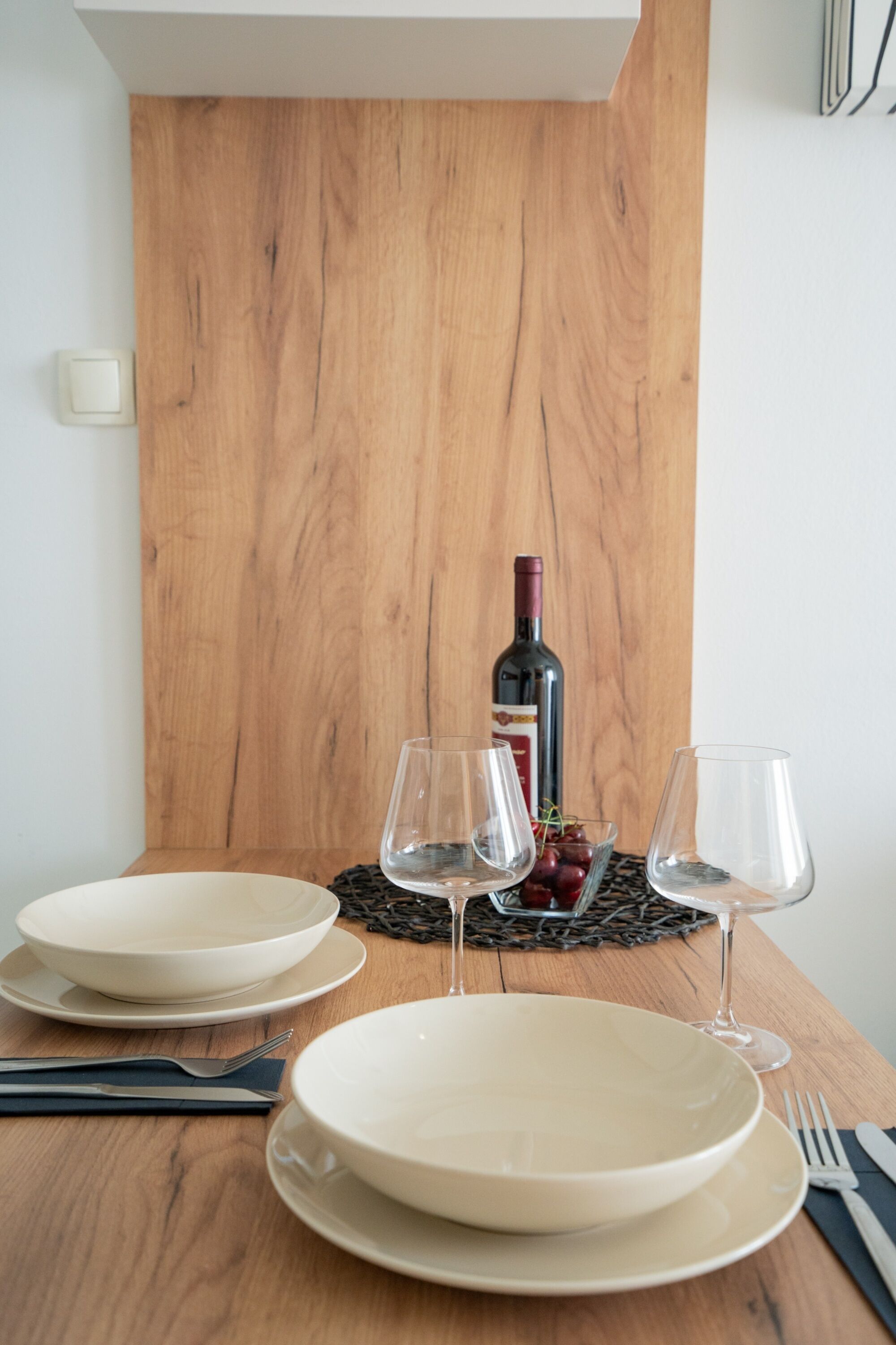 A dining table set with plates, glasses, and cutlery, with a bottle of wine and grapes on a placemat.