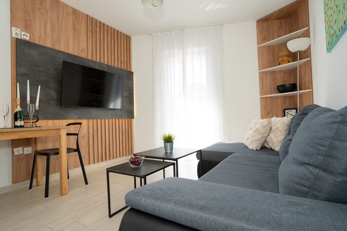 A modern living room with a grey sofa, wooden panel walls, a flat-screen TV, a dining area with a wooden table and chairs, and a small side table with a plant.