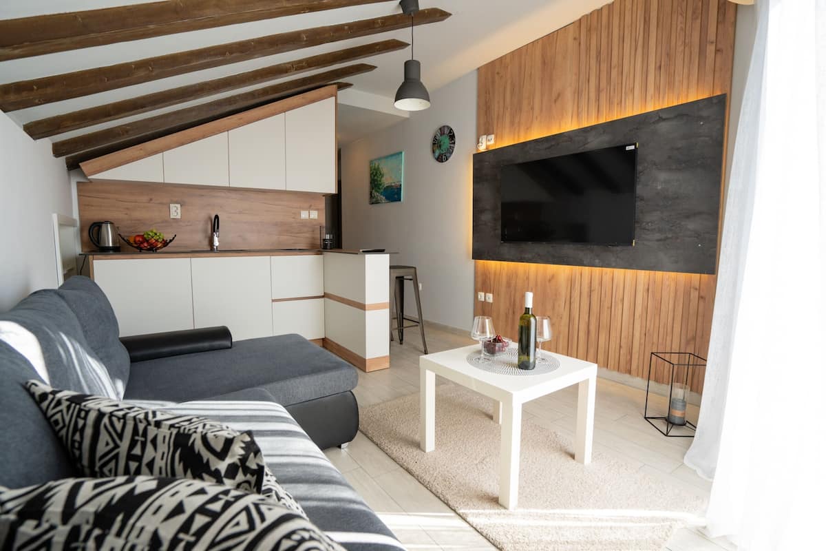 A modern living room with a grey sofa, a white coffee table, and a kitchen area with wooden cabinets and a black TV mounted on the wall.