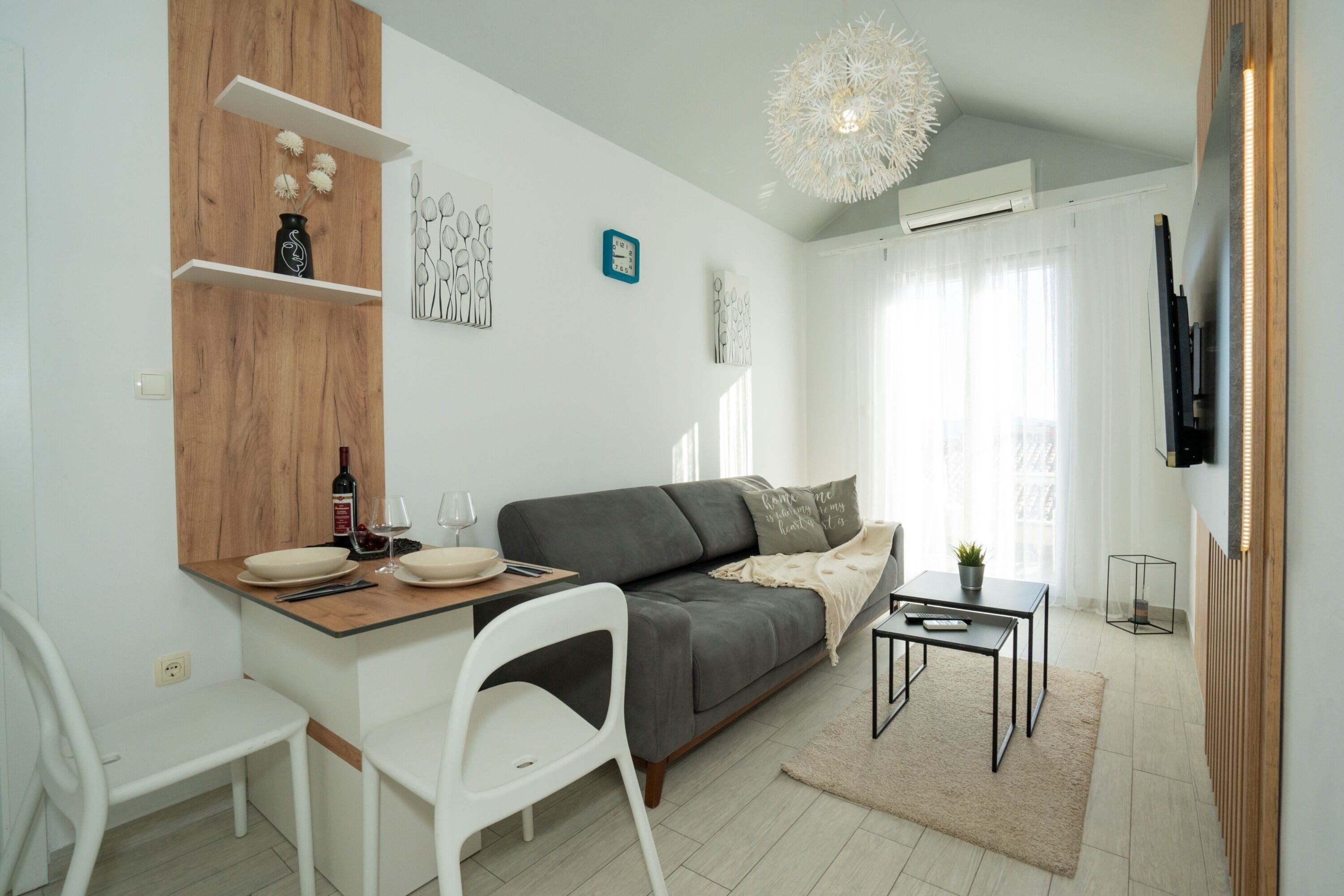 A modern living room with a grey sofa, a wooden accent wall, a dining table with chairs, and a TV mounted on the wall.