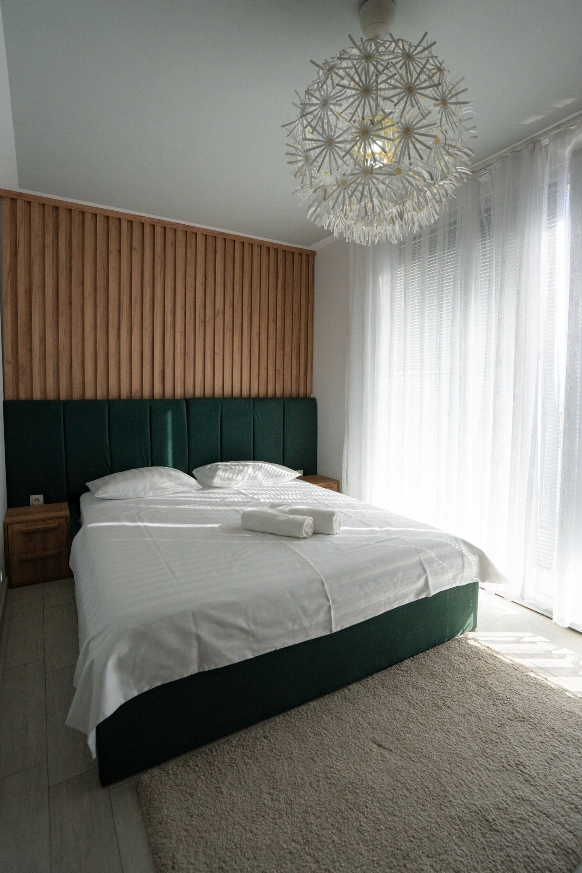 Un dormitorio con cama, cabecera de madera, ropa de cama blanca, alfombra blanca y lámpara colgante.
