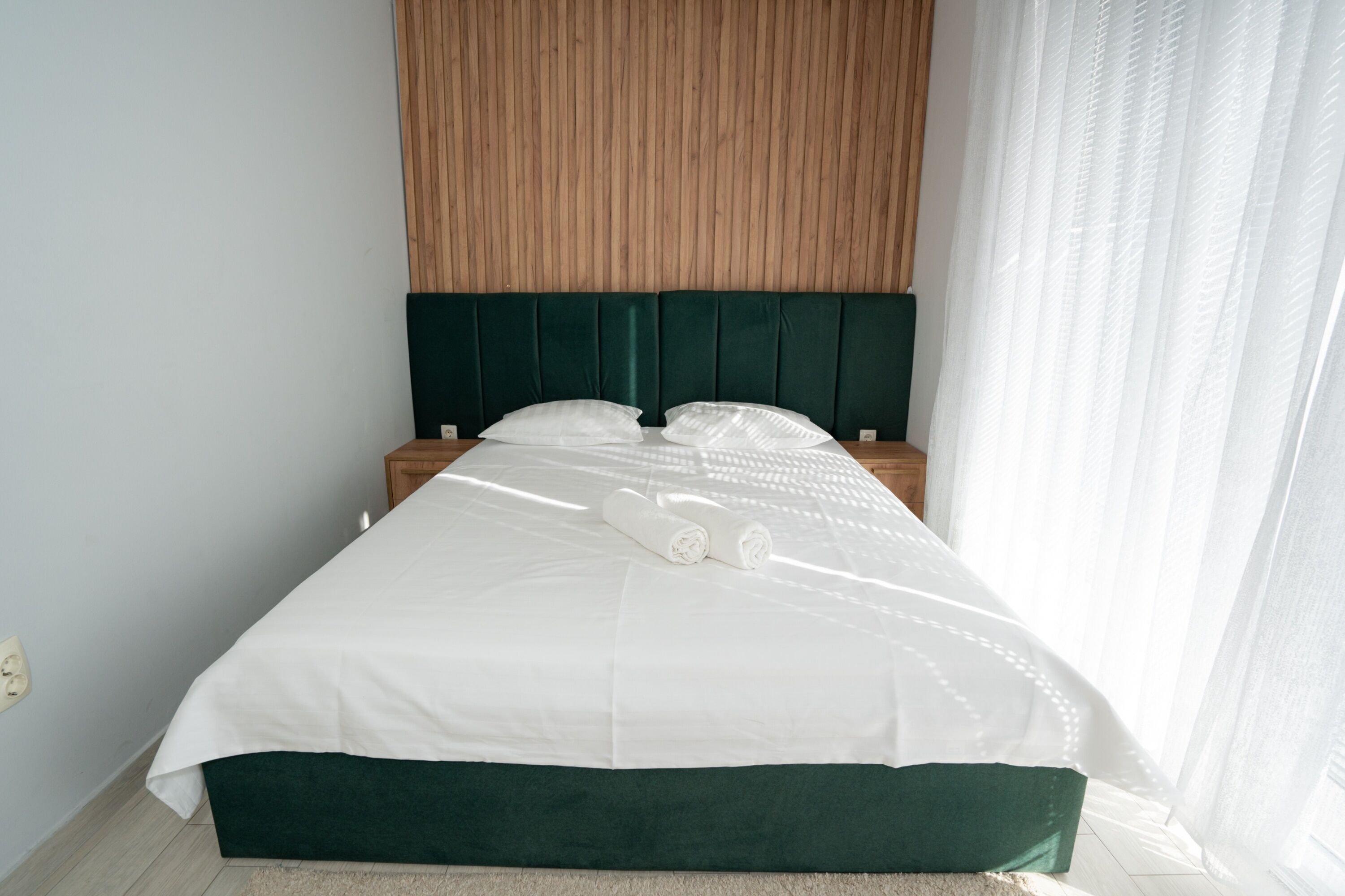A neatly made bed with a green headboard, white bedding, and two rolled towels.