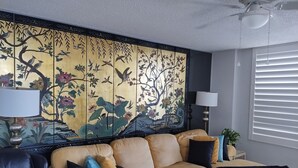 En stue med en beige sofa, sorte og blå puder, en dekorativ vægpanel med fugle og blomster, en ventilator i loftet og et vindue med persienner.