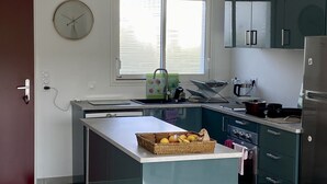 A modern kitchen with a central island, a sink, a stove, and a refrigerator. There is a window with blinds, a clock on the wall, and a door to the left.