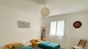 A room with two single beds, a wooden chair, a small table, a bookshelf, and a decorative wall piece.