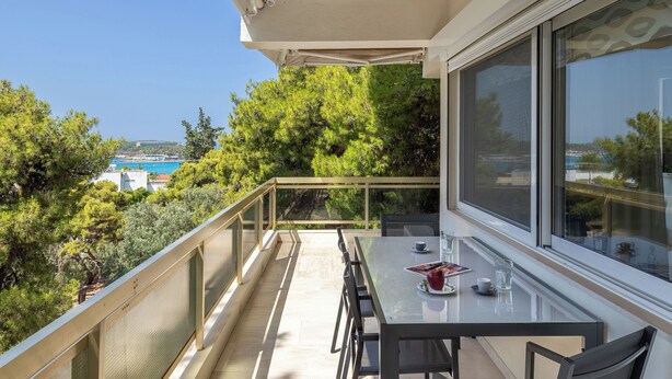 A balcony with a table and chairs, overlooking a scenic view of trees and a body of water. - Relux Sea View Two Bedroom Apartment by BluPine (Athens)
