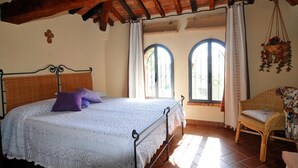 Une chambre à coucher avec un lit, deux fenêtres, un fauteuil en osier et une plante suspendue.
