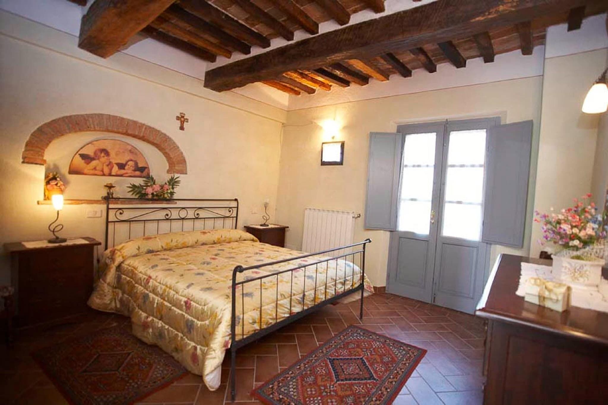 A bedroom with a bed, wooden ceiling beams, a window with shutters, a radiator, and a small table with a vase of flowers.