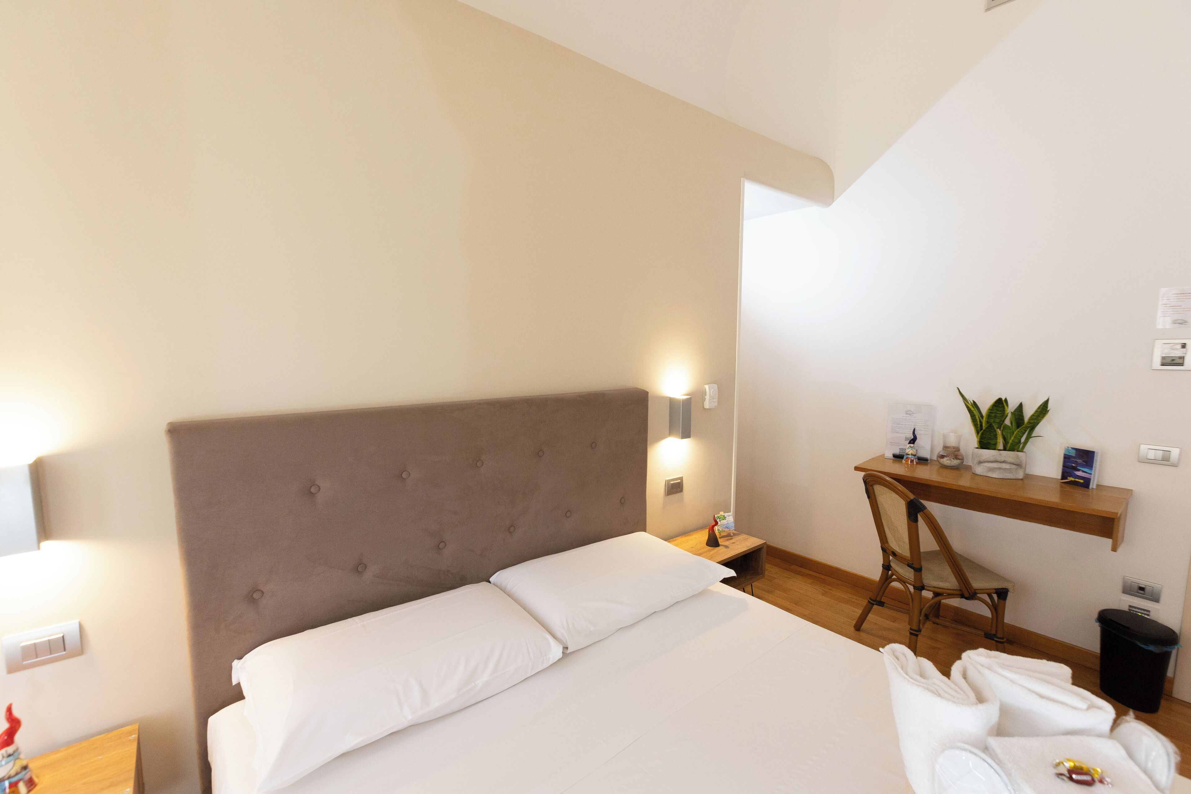 Ein Schlafzimmer mit einem großen Bett, einem Nachttisch, einem Sessel, einem Schreibtisch mit Pflanze und einem Papierkorb.