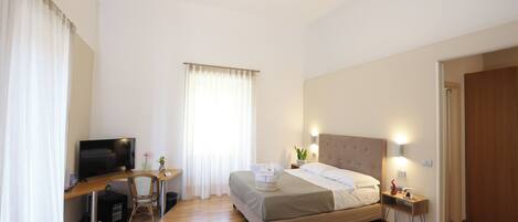 Habitación de hotel con cama, escritorio, computadora, televisor y mesita de noche con lámpara.