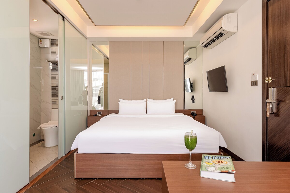 Una habitación de hotel con una cama grande, una mesita de noche con un libro y un vaso de bebida verde, un televisor montado en la pared y un equipo de aire acondicionado.