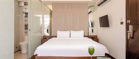 Hotelkamer met een groot bed, nachtkastje met een boek en een glas groene drank, een aan de muur bevestigde televisie en een airconditioning.