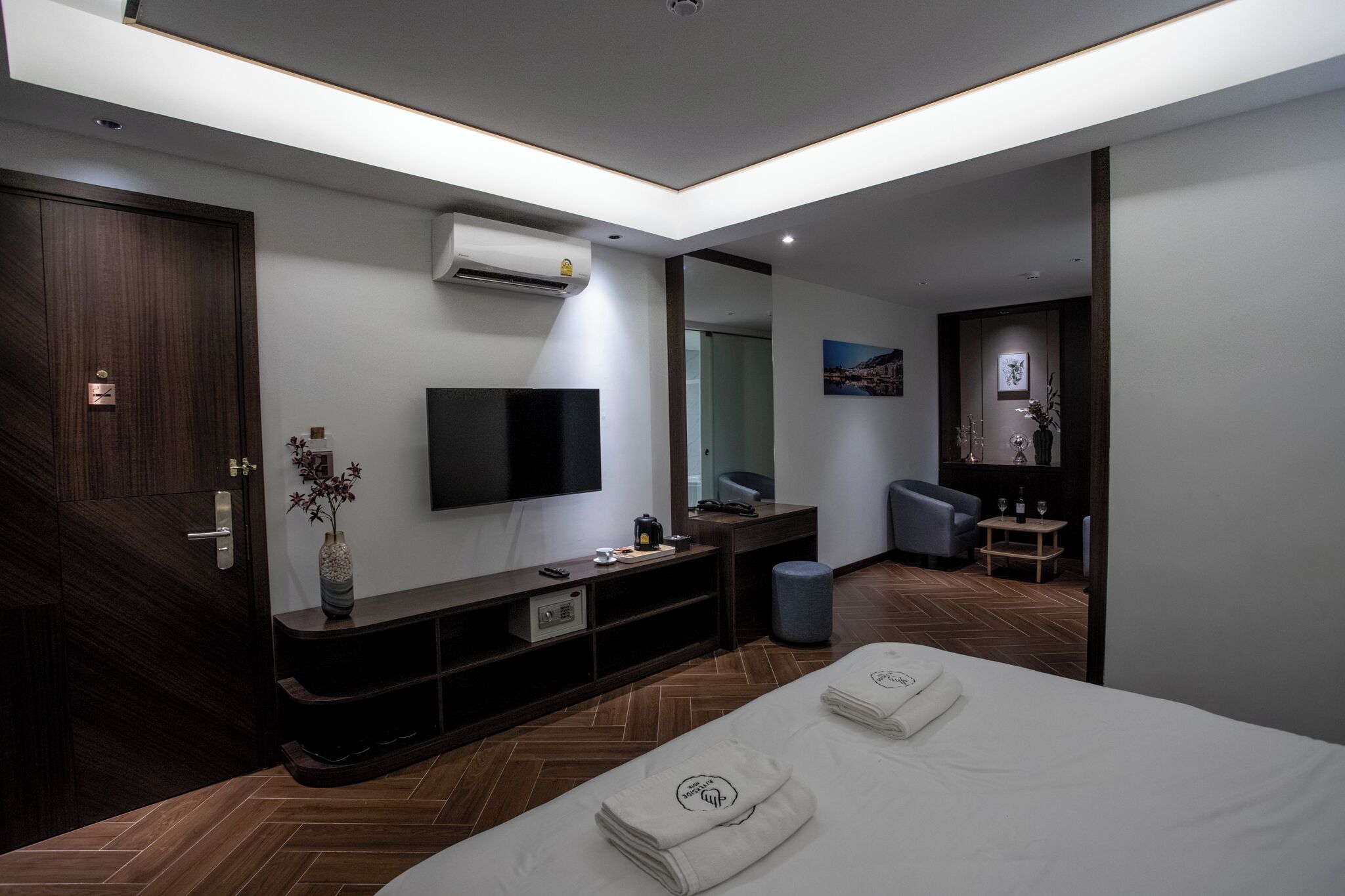 A modern hotel room with a large bed, a flat-screen TV mounted on the wall, a small seating area with a chair and table, and a wooden floor.