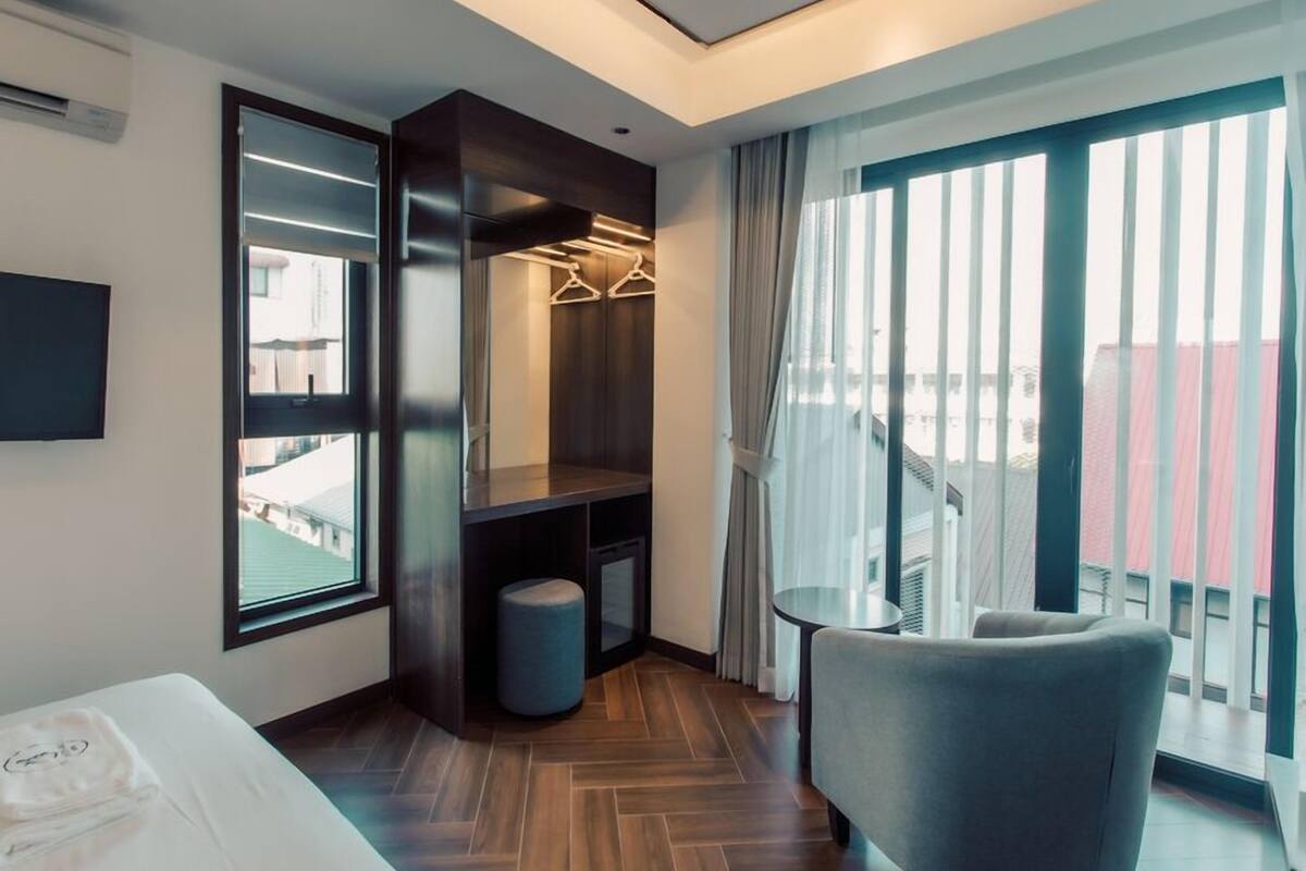 A modern hotel room with a large window, a seating area, a wardrobe, and a small trash bin.