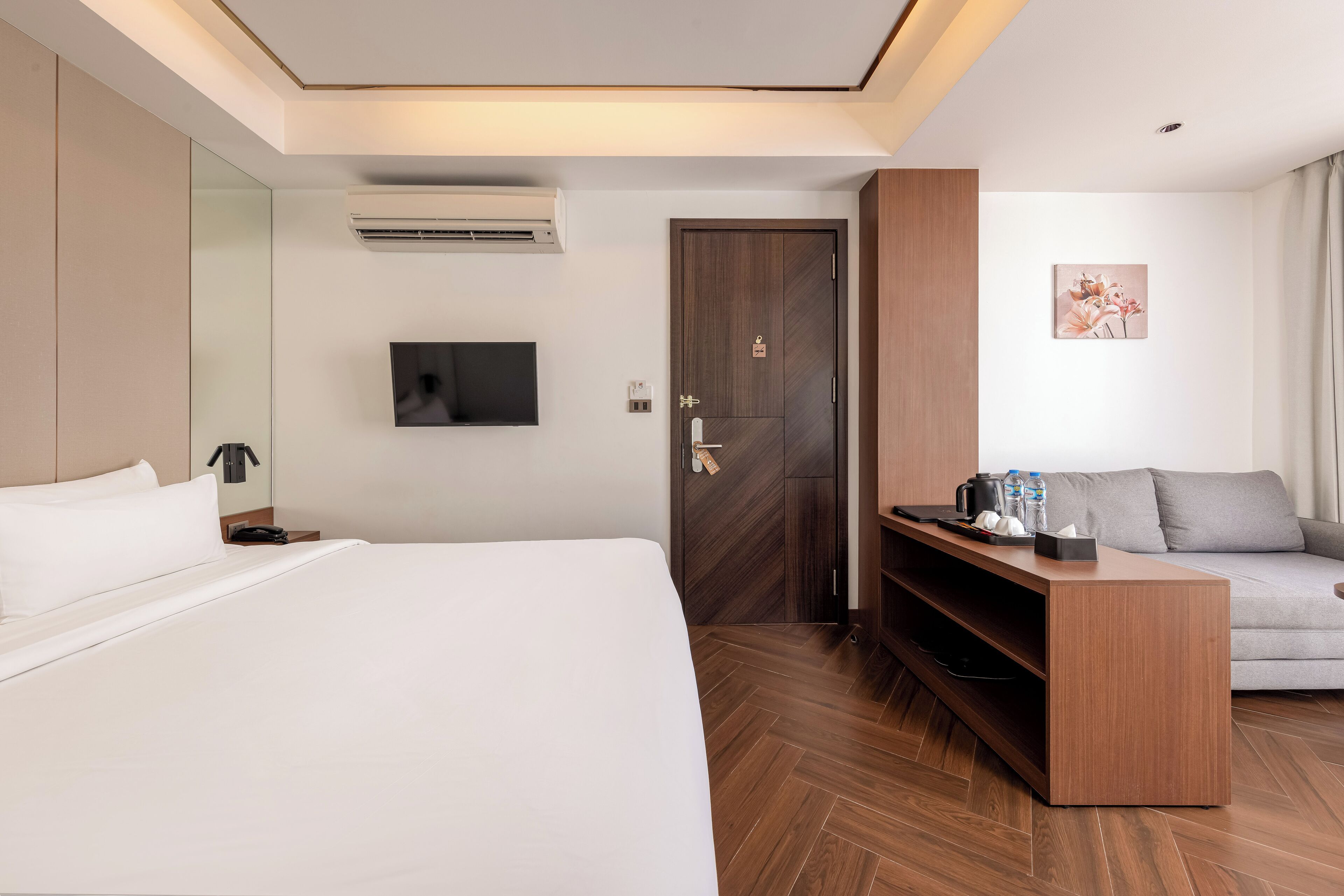A hotel room with a bed, a TV mounted on the wall, a wooden door, a grey sofa, and a wooden console table with items on it.