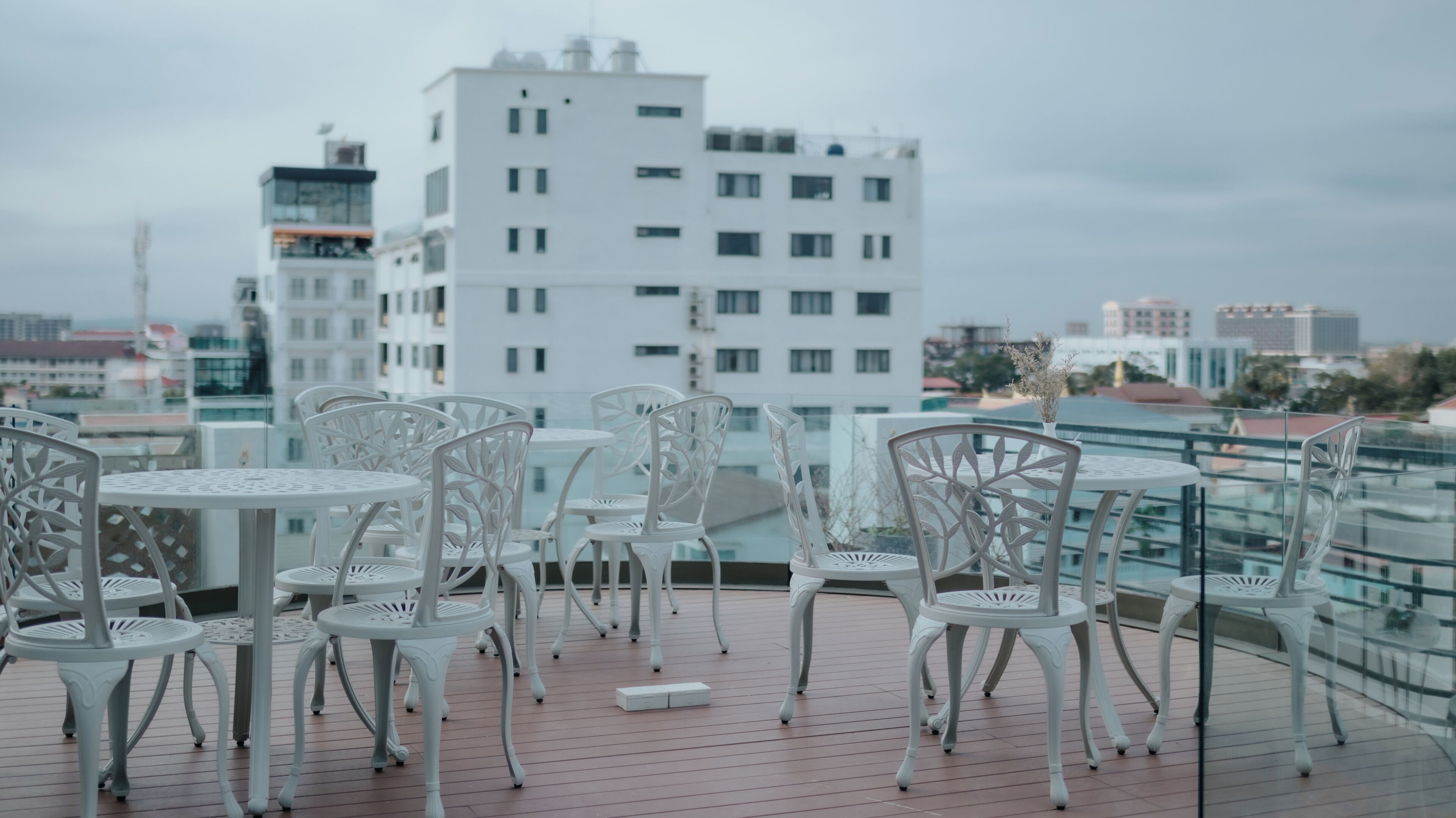 Terraza en la azotea con sillas y mesas de metal blanco, con vistas a un paisaje urbano de edificios modernos.