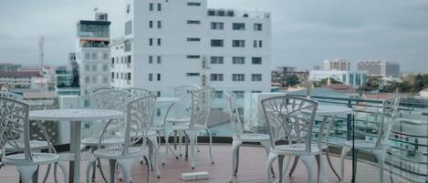 Terraza en la azotea con sillas y mesas de metal blanco, con vistas a un paisaje urbano de edificios modernos.