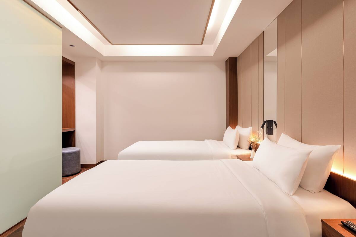 A hotel room with two beds, a bedside table with a lamp, and a small seating area with a cushion.