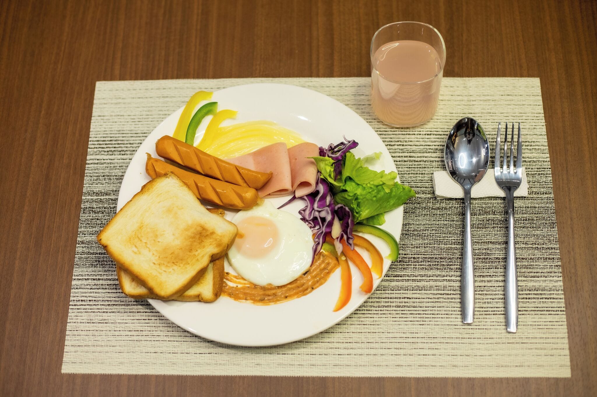 A plate with toast, a fried egg, ham, and vegetables, accompanied by a glass of water and cutlery on a placemat.