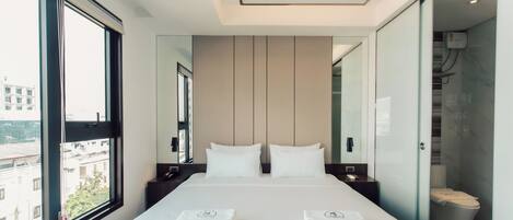 A modern hotel room with a large bed, two bedside tables, a mirror, and a view of the city through a large window.