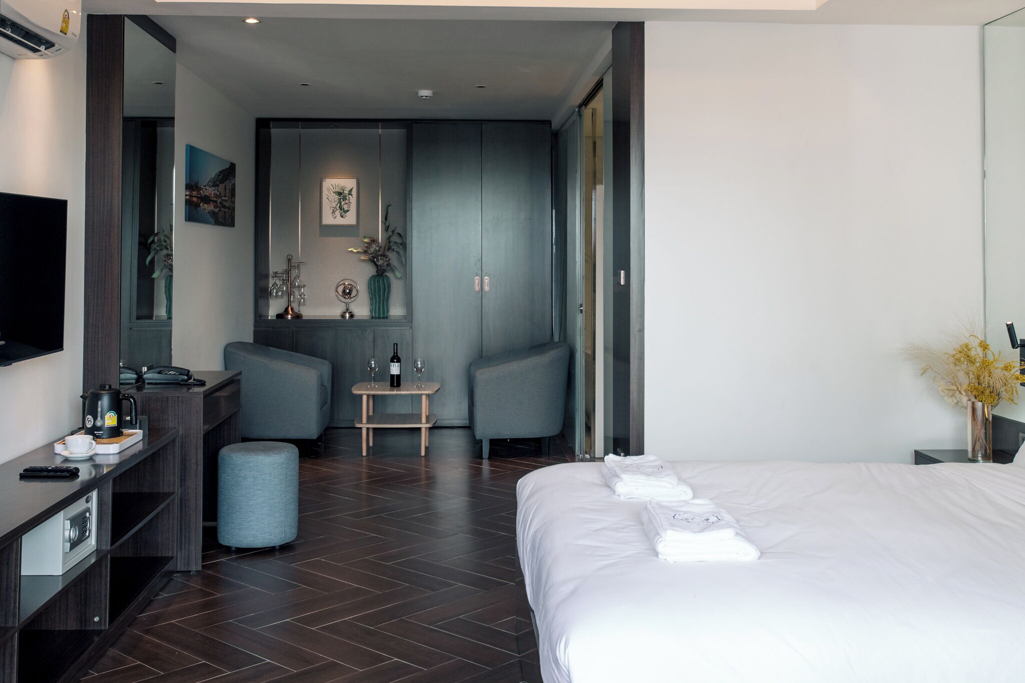 A modern hotel room with a large bed, a desk with a computer, a chair, a small table with a vase and wine glasses, and a large mirror.