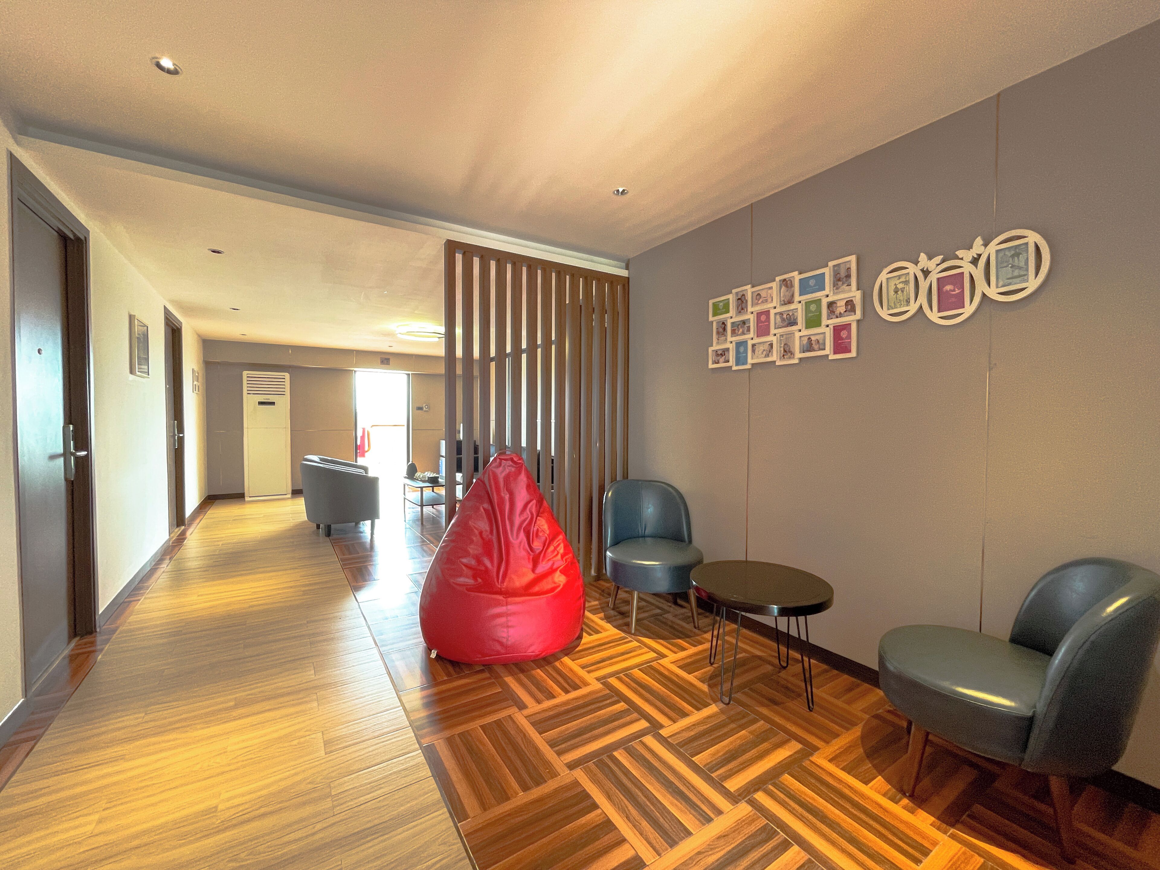 Un hall d’entrée moderne avec un parquet en bois disposé en chevrons, un fauteuil-pouf rouge, deux fauteuils gris et un mur décoré de photos encadrées.
