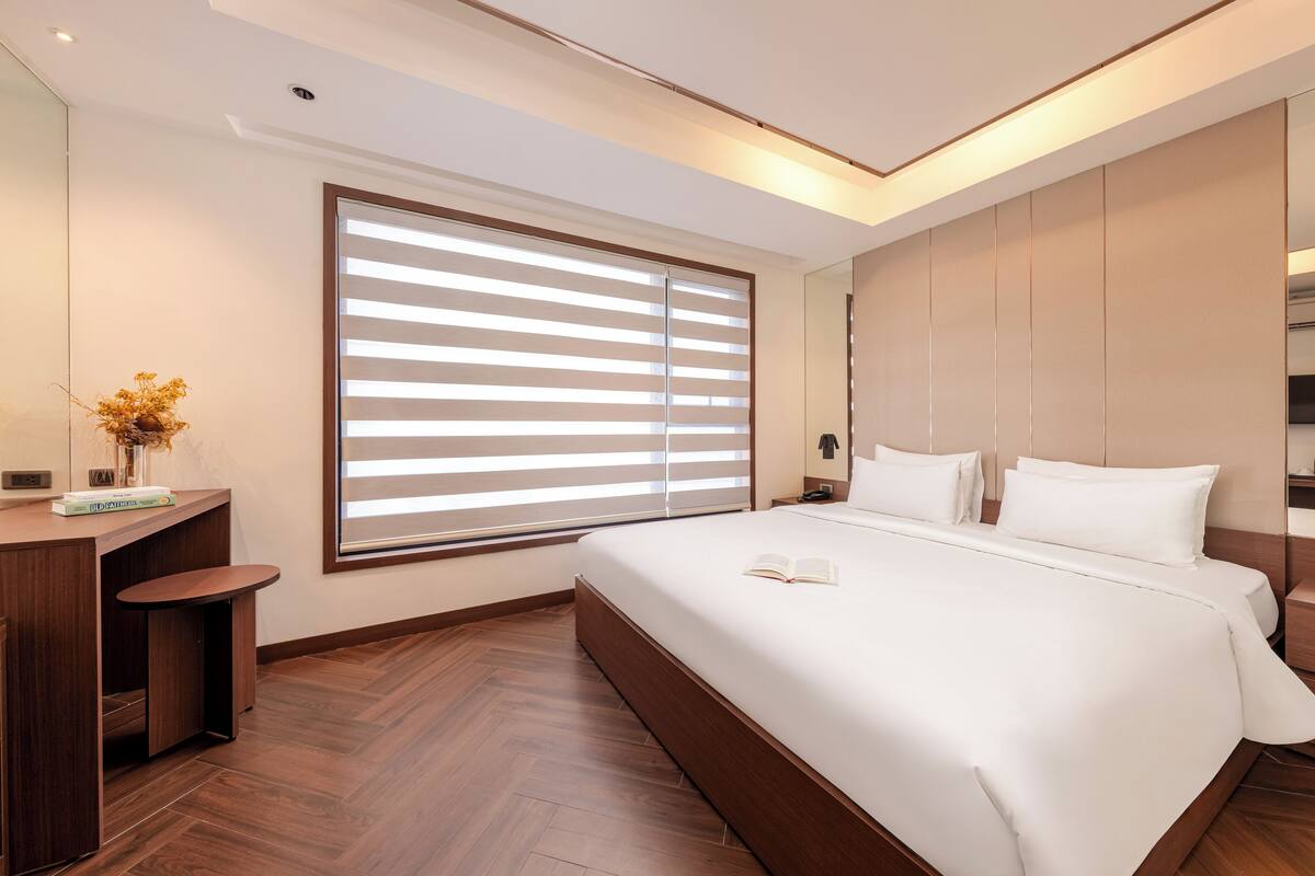 A modern hotel room with a large bed, a bedside table, a desk, and a large window with blinds.