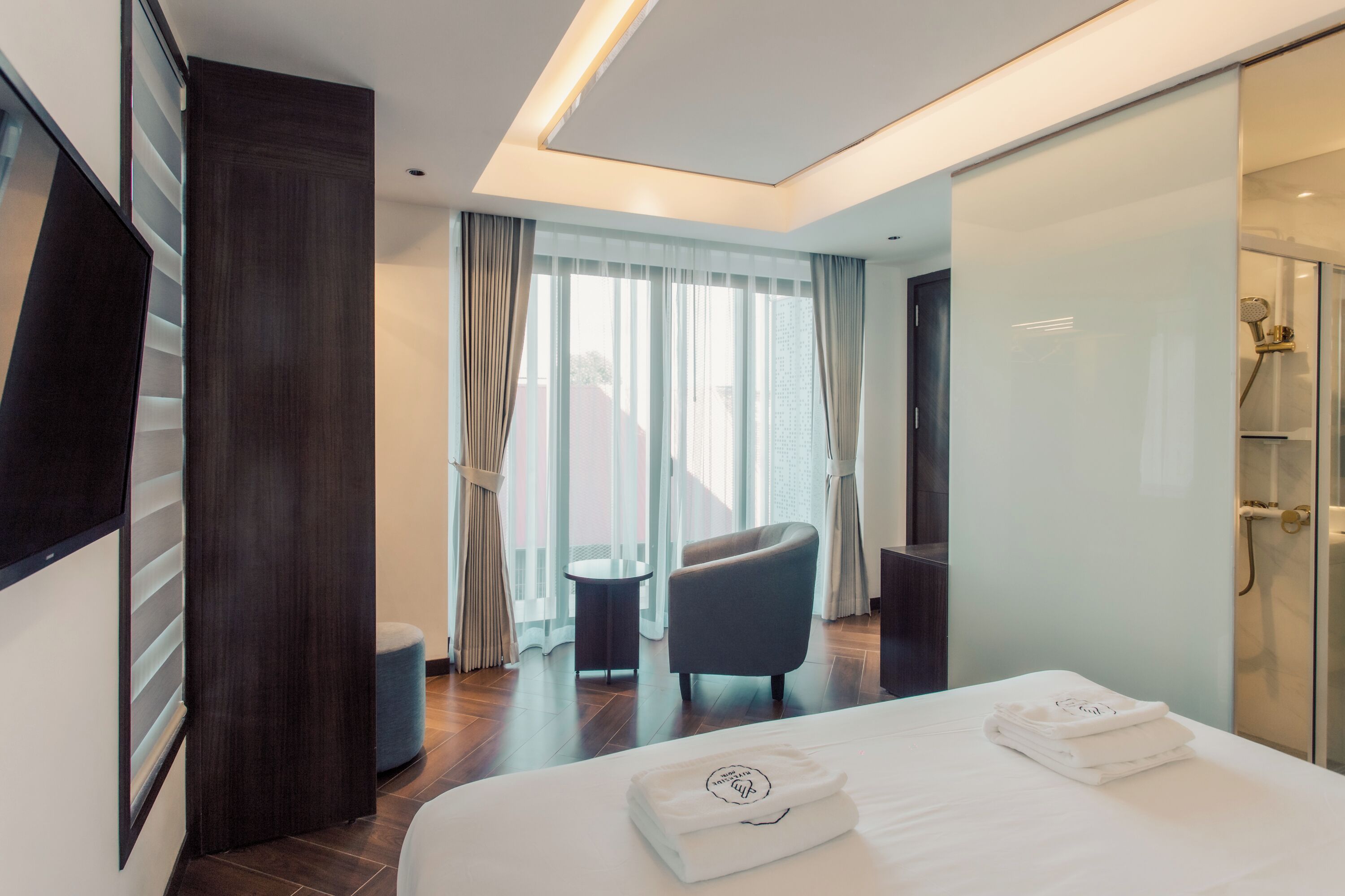 A modern hotel room with a large bed, a flat-screen TV, a seating area with a chair, and a glass-enclosed shower.