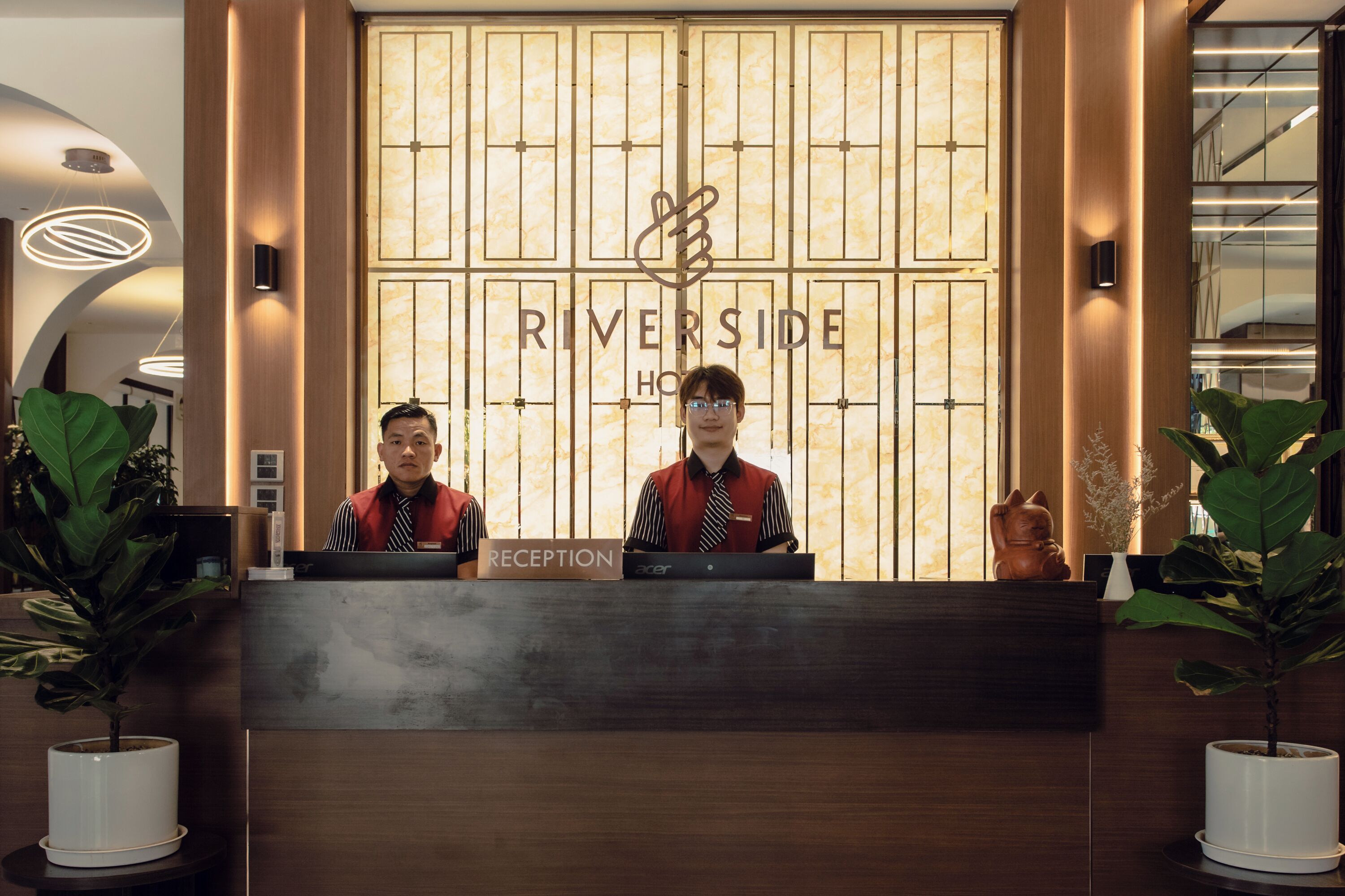A hotel reception desk with two staff members, a reception sign, and a large window with a decorative design.