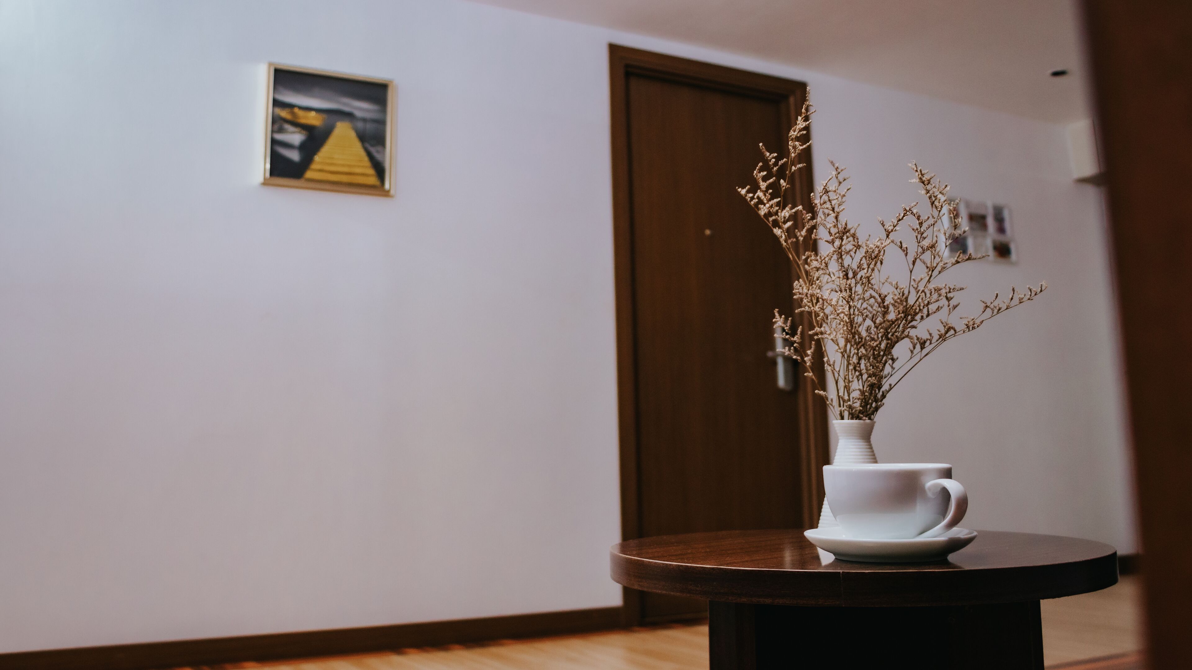 A hallway with a wooden floor, a white vase with dried flowers on a round table, and a door in the background.